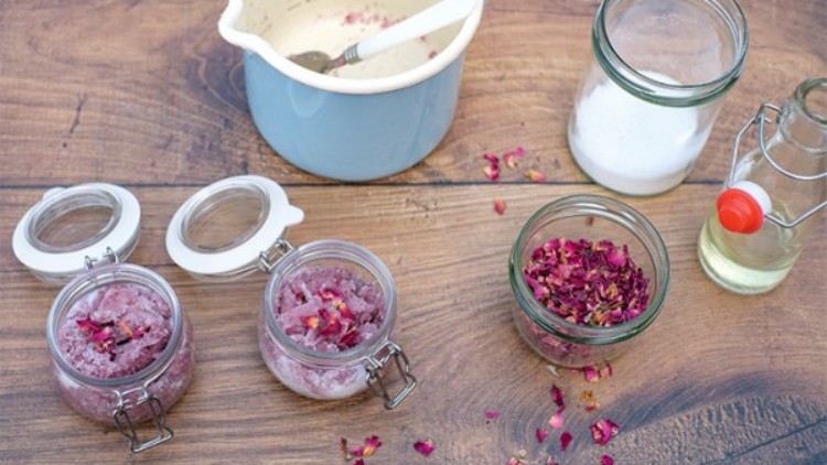 Mehrere Einmachgläser mit rosafarbenem Zucker und Rosenblüten, eine blaue Rührschüssel, Salz und eine Glasflasche auf Holztisch.