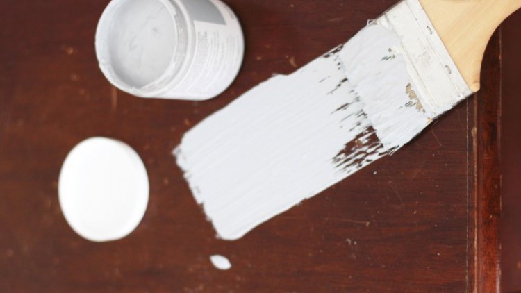 Ein breiter Pinsel mit weißer Farbe liegt auf einer dunklen Holzoberfläche, daneben ein offener Farbtopf mit Deckel.