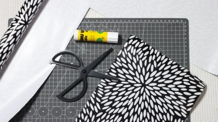 Schwarze Schere, Klebestift und schwarz-weiß gemustertes Papier liegen auf einer Schneidematte mit Rasterlinien und Kreisen.