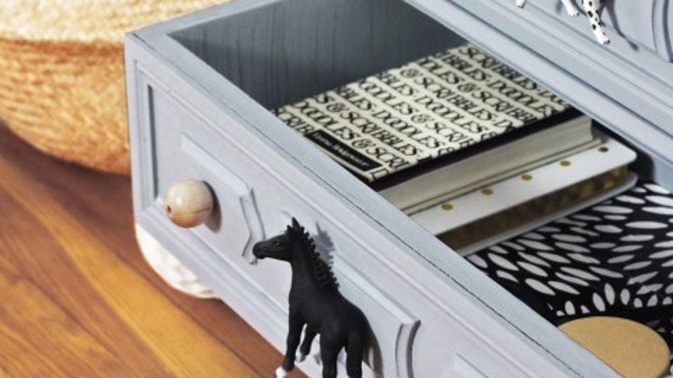 Geöffnete graue Holzschubladen mit Tierfiguren als Griffe, daneben Körbe mit schwarzen Luftballons und gemusterten Accessoires.