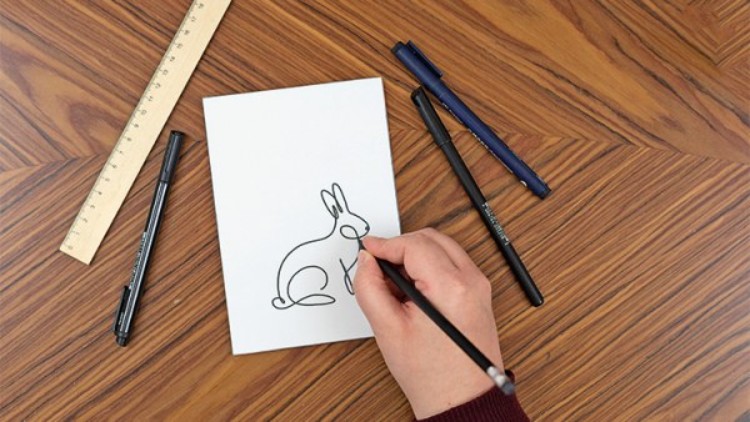 Eine Hand zeichnet mit einem schwarzen Stift einen Hasen auf weißen Karton, daneben liegen Stifte und ein Lineal auf Holztisch.