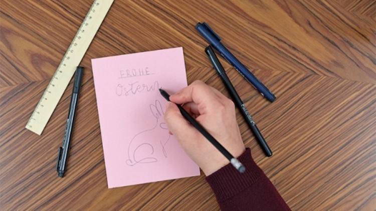 Eine Hand zeichnet mit einem schwarzen Stift einen Hasen auf eine rosa Karte mit der Aufschrift Frohe Ostern, umgeben von Stiften.