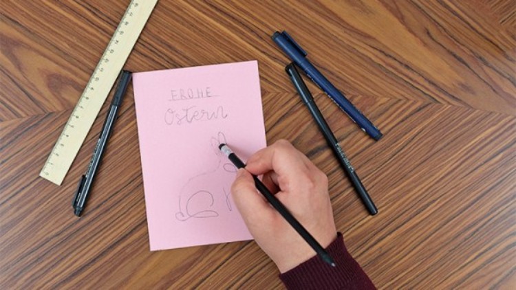 Eine Hand zeichnet mit einem schwarzen Stift einen Hasen auf eine rosa Karte mit der Aufschrift Frohe Ostern, umgeben von Stiften.