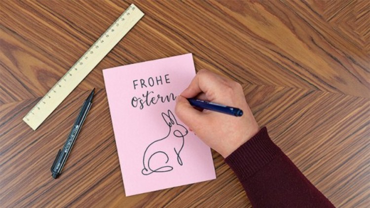 Eine Hand zeichnet mit einem blauen Stift ein Kaninchen auf eine rosa Karte mit der Aufschrift Frohe Ostern, daneben Lineal und Stift.