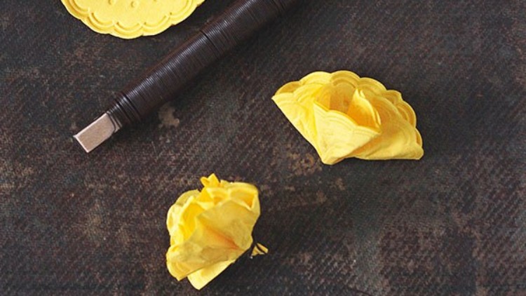 Gelbes Bastelpapier, teilweise zu Blumen geformt, liegt neben einer dunklen Rolle mit hellem Griff auf einer dunklen Oberfläche.