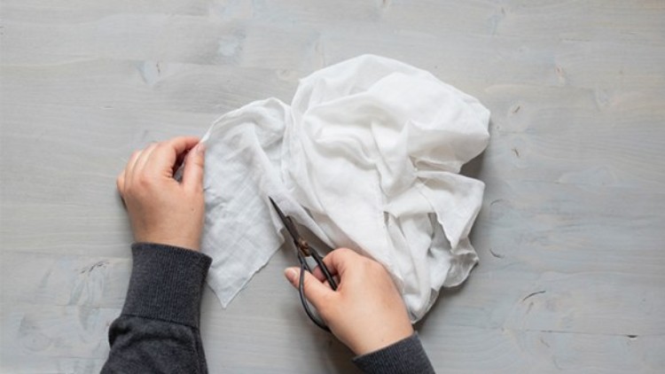 Zwei Hände schneiden mit einer Schere ein Stück weißen Stoff auf einem hellgrauen Holzuntergrund zurecht.