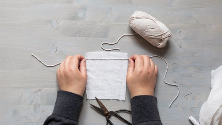 Zwei Hände falten weißen Stoff, daneben liegen weißes Garn, eine Schere und ein Holzuntergrund im grauen Farbton.