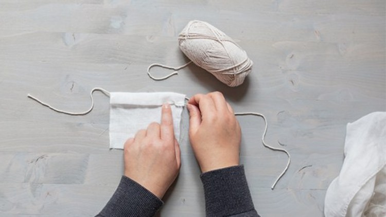 Zwei Hände nähen mit weißem Garn an einem weißen Stoffstück, daneben liegt ein Knäuel weißen Garns auf hellem Untergrund.