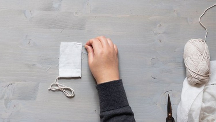 Eine Hand näht mit einer Nadel einen weißen Stoffbeutel, daneben liegen weißes Garn, Stoff und eine Schere.