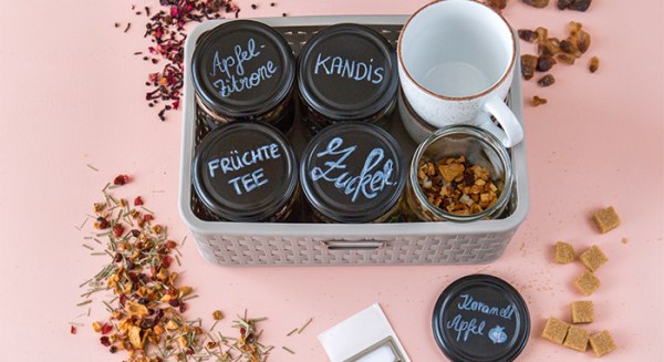 Grauer Korb mit Teegläsern, Kandis, Zucker, einer Tasse und Streutee, Deckel mit Beschriftungen wie Apfel-Zitrone und Früchte-Tee.