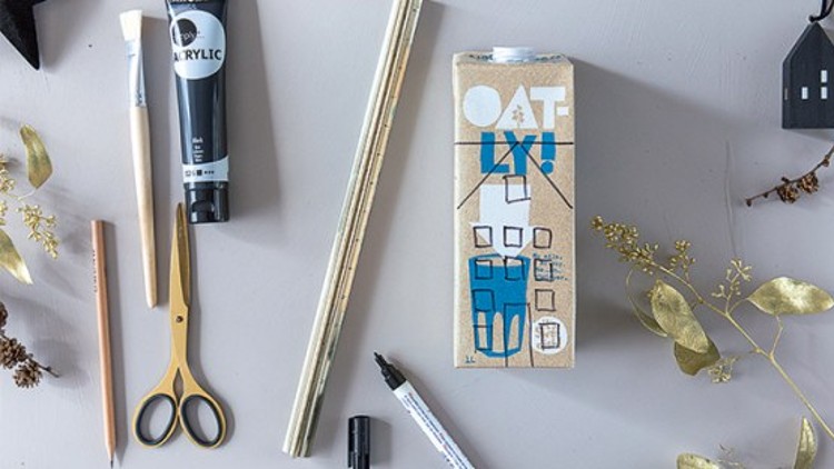 Ein Karton mit der Aufschrift „OATLY“ steht auf einem Tisch, umgeben von Stiften, Schere, Farbe, Pinseln und goldenen Deko-Elementen.