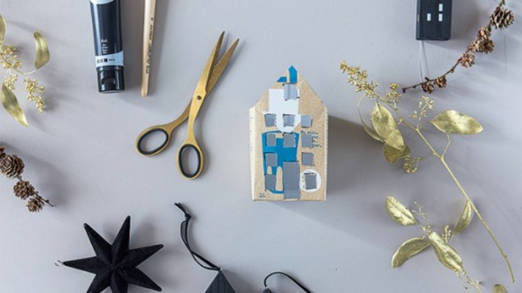 Kleines, bastelhaftes Haus aus Pappe mit blauen und weißen Farbfeldern, umgeben von Schere, Farbe, goldenen Zweigen und Dekoration.