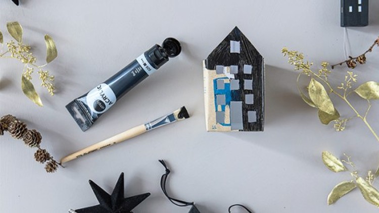 Kleines bemaltes Holzhaus, schwarze Acrylfarbe mit Pinsel, goldene Zweige und schwarze Dekorationen auf grauem Untergrund.