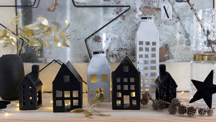 Dekorative Miniaturhäuser in Schwarz und Weiß mit Lichterkette, neben Zapfen, goldenen Zweigen und einem schwarzen Stern.