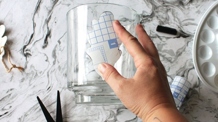 Eine Hand hält eine selbstklebende Folie mit blauem Karomuster, die auf ein transparentes Glasgefäß geklebt wird.