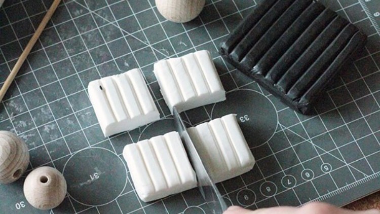 Eine Hand schneidet weiße Modelliermasse mit einem Messer, daneben liegt schwarze Modelliermasse und Holzkugeln auf einer Schneidematte.