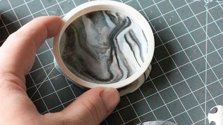 Eine Hand hält eine flache runde Schale mit grauen und weißen Marmormustern auf einer Schneidematte mit Rastermuster.