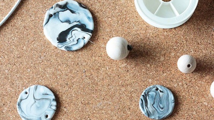 Mehrere flache Anhänger mit Marmormuster in Schwarz-Weiß und Holzperlen liegen auf einer Korkunterlage, daneben eine weiße Garnspule.