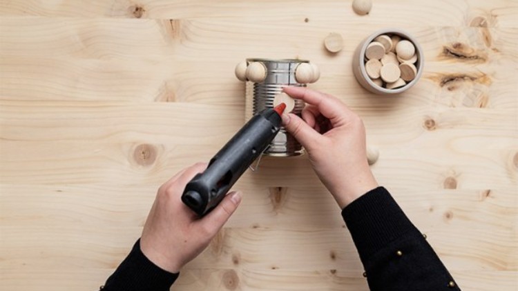 Zwei Hände befestigen mit einer Heißklebepistole Holzperlen auf einer Konservendose, daneben liegt ein Gefäß mit Holzscheiben.