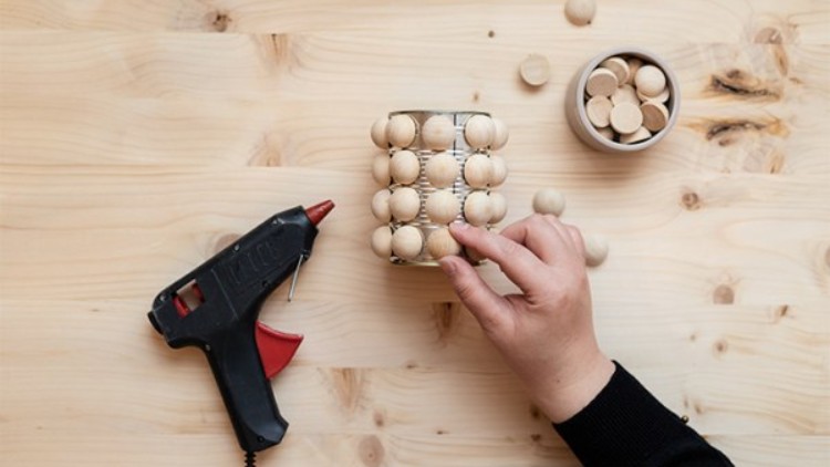 Hand fixiert Holzperlen mit einer Heißklebepistole an einem Glas, daneben eine Schale mit weiteren Holzperlen auf hellgemasertem Holz.