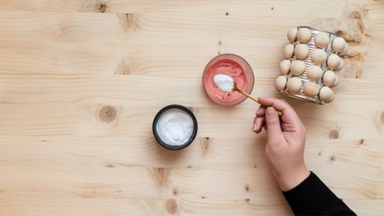 Eine Hand hält einen kleinen goldfarbenen Löffel mit weißer Creme über einer offenen Schale mit roter Creme auf Holztisch.