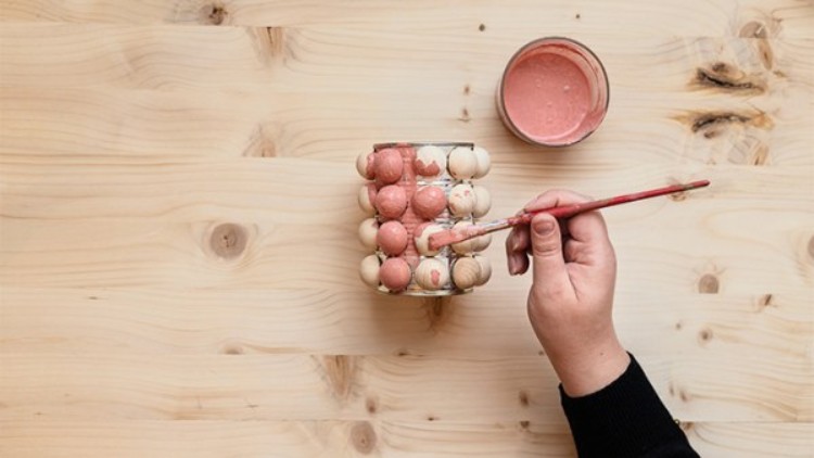 Hand bemalt weiße und roséfarbene Holzkugeln auf einem Glas, daneben steht ein offenes Gefäß mit rosafarbener Farbe.