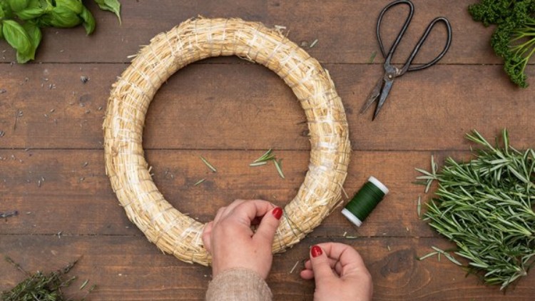 Eine Person bindet grüne Zweige mit einem Draht an einen Strohring, auf einem Holztisch liegen Kräuter und eine Schere.