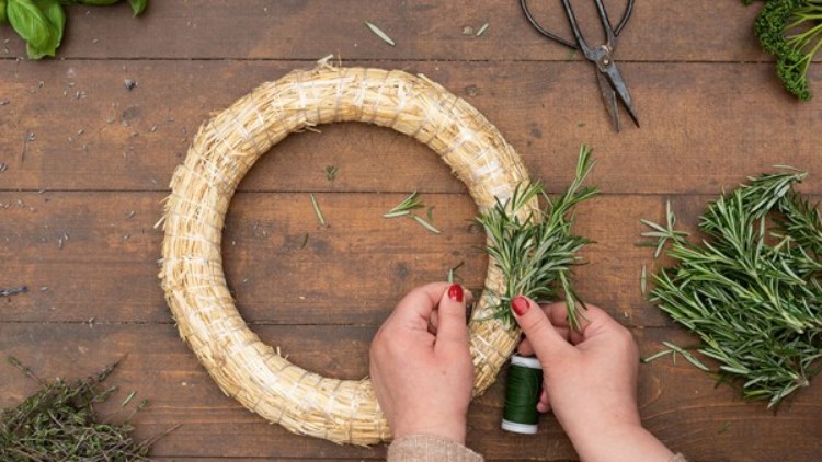 Eine Person bindet Rosmarinzweige mit grünem Draht an einen Strohring, umgeben von Kräutern, Schere liegt daneben.