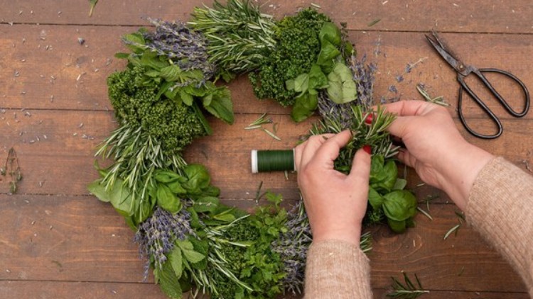 Zwei Hände binden mit grünem Faden einen Kräuterkranz aus Rosmarin, Lavendel, Basilikum und weiteren Kräutern auf Holztisch.