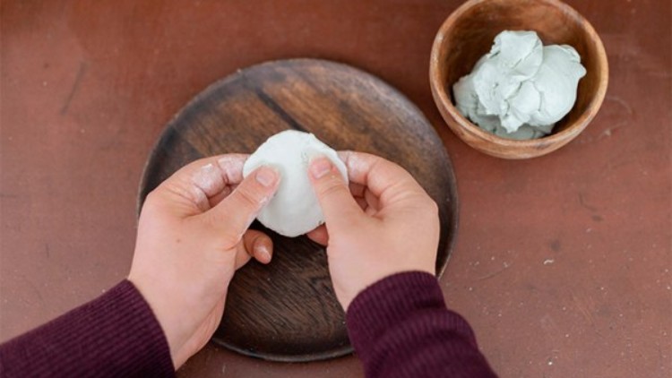 Zwei Hände formen weißen Teig auf einem braunen Teller, daneben eine Holzschale mit weiterer Teigmasse.