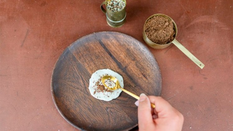 Eine Hand hält einen goldfarbenen Löffel und gibt braunes Pulver auf weiße Creme auf einem braunen Holzteller.