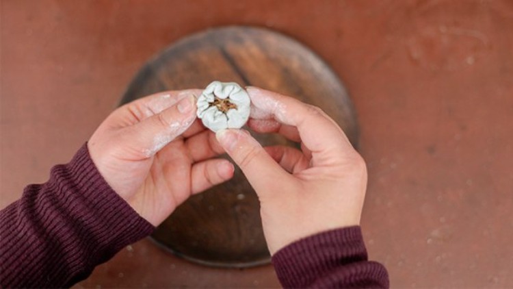 Zwei Hände in einem weinroten Oberteil halten einen weißen Teigling mit gefaltetem Rand und einer geöffneten Mitte.