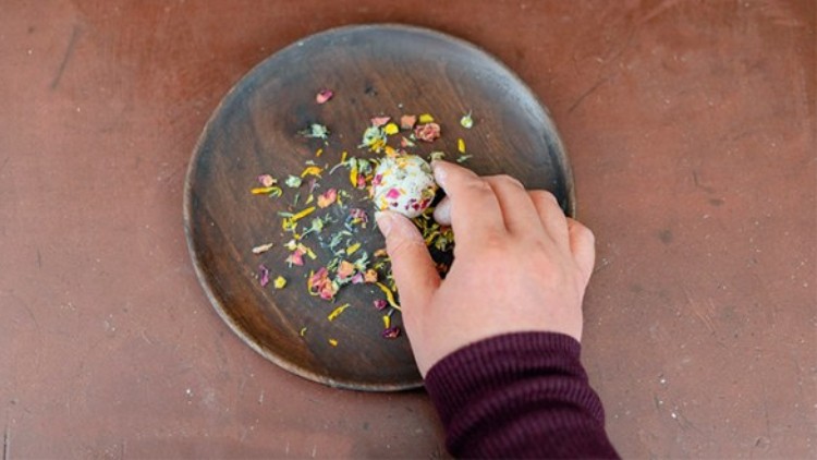 Hand greift nach einer bunt verzierten Praline auf einem dunklen Teller mit getrockneten Blüten auf braunem Untergrund.