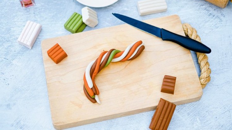 Verschiedene Knetblöcke und ein schwarzgriffiges Messer liegen auf einem Holzbrett mit gedrehter Knetfigur in Grün, Weiß und Orange.