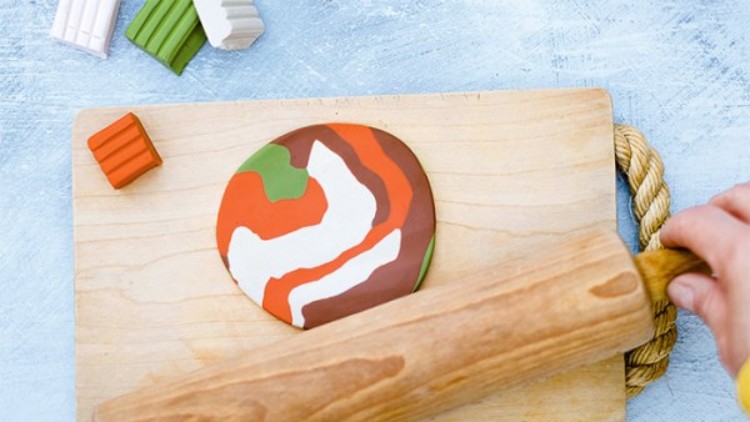 Eine Hand rollt bunten Knetmasse-Teig in Braun, Grün, Weiß und Orange mit einem Holzroller auf einem Holzbrett aus.