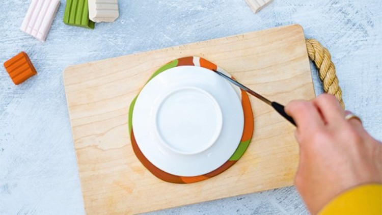 Eine Hand schneidet mit einem Messer um eine weiße Schale auf farbiger Modelliermasse auf einem Holzbrett.