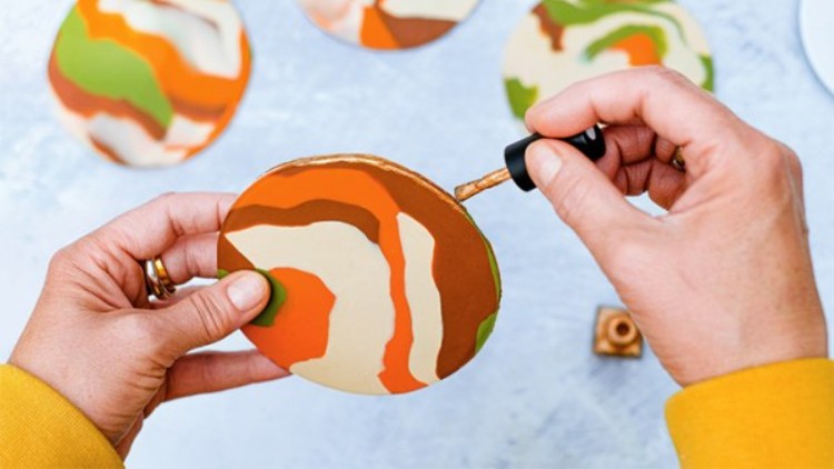 Hände halten einen bemalten runden Untersetzer, während mit einem Pinsel eine Linie in Orange hinzugefügt wird.