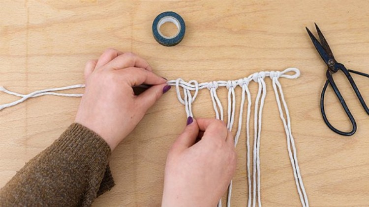 Hände knoten weiße Schnüre auf einem Holztisch, daneben liegen eine Schere und eine Rolle schwarzes Klebeband.
