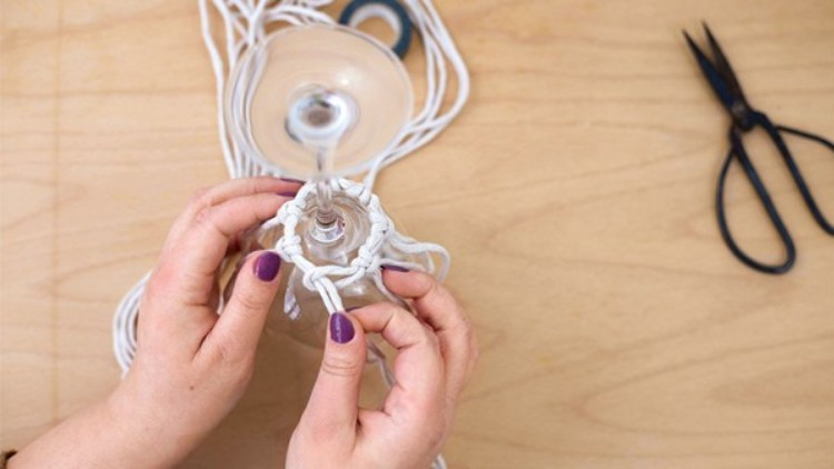 Hände mit lila lackierten Nägeln knoten weiße Schnüre um den Stiel eines umgedrehten Glases, daneben liegt eine Schere.