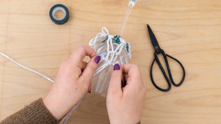 Eine Person knotet weiße Schnur an den Stiel eines Weinglases, daneben liegen Schere und Klebeband auf holzfarbenem Untergrund.