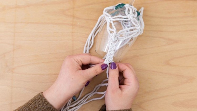 Zwei Hände mit lila lackierten Nägeln flechten weiße Schnüre um einen Plastikbecher auf einer Holzoberfläche.