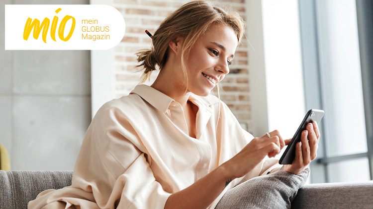 Person in beiger Bluse sitzt auf einem Sofa und nutzt ein Smartphone, Logo „mio mein GLOBUS Magazin“ ist oben links zu sehen.