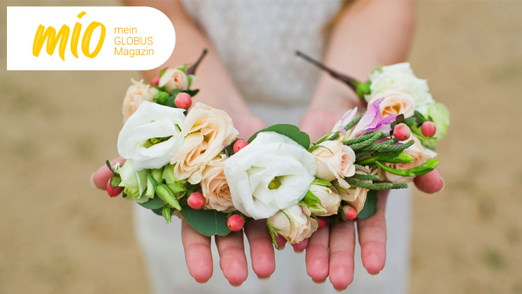 Hände halten einen Blumenkranz mit weißen, pinken und beigen Blüten sowie grünen Blättern, links steht „mio mein GLOBUS Magazin“.
