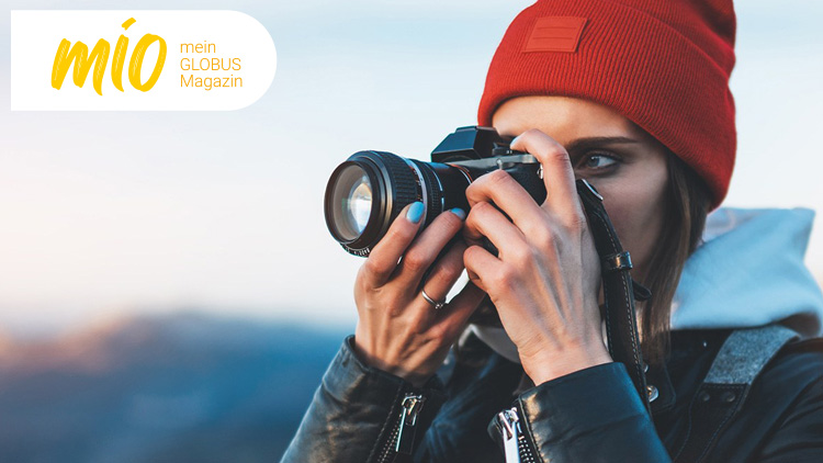 Person in outdoor Kleidung hält eine Kamera in den Händen, oben links steht mio mein GLOBUS Magazin in gelber Schrift.