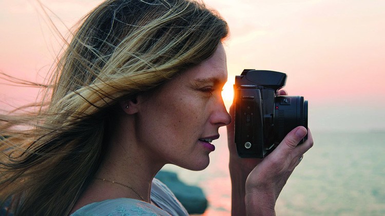Eine Person hält eine Kamera vor das Gesicht, im Hintergrund Sonnenuntergang über einem Gewässer.