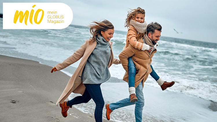 Drei Personen in warmer Kleidung laufen lachend am Strand entlang, im Hintergrund das Meer und eine fliegende Möwe.