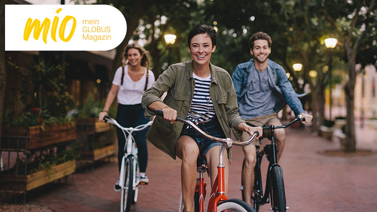 Drei Personen in Freizeitkleidung fahren auf Fahrrädern durch eine beleuchtete Allee, oben links steht „mio mein GLOBUS Magazin“.