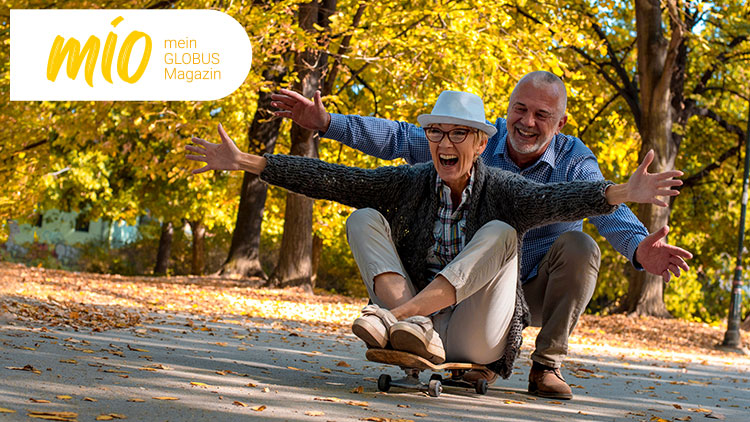 Zwei Personen fahren auf einem Skateboard durch einen herbstlichen Park, links oben steht „mio mein GLOBUS Magazin“.