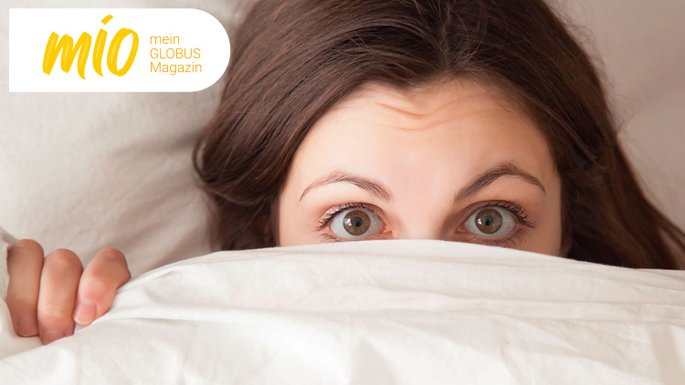 Eine Hand hält eine weiße Bettdecke, im Hintergrund braune Haare, links oben steht „mio mein GLOBUS Magazin“ in Gelb und Weiß.