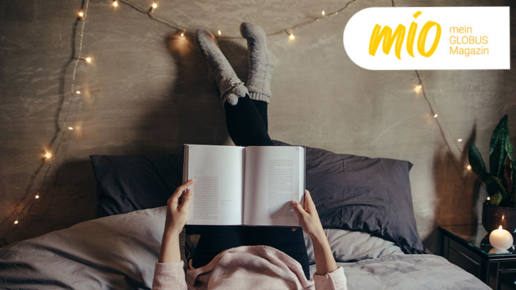 Person liegt auf einem Bett mit grauer Bettwäsche, liest ein Buch und rechts oben steht „mio, mein GLOBUS Magazin“.